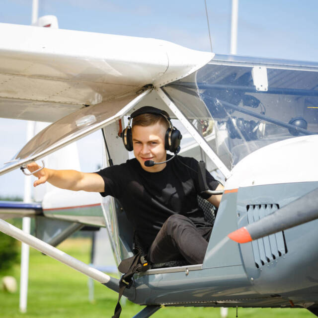 handsome teenage man sitting in small plane cockpit. young pilot with headset on. outdoor shot.